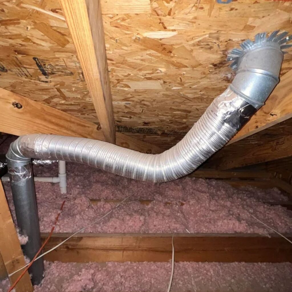 A silver metal dryer vent transition pipe in the attic of a home. There is silver metal duct tape on the pipe where it has been repaired.